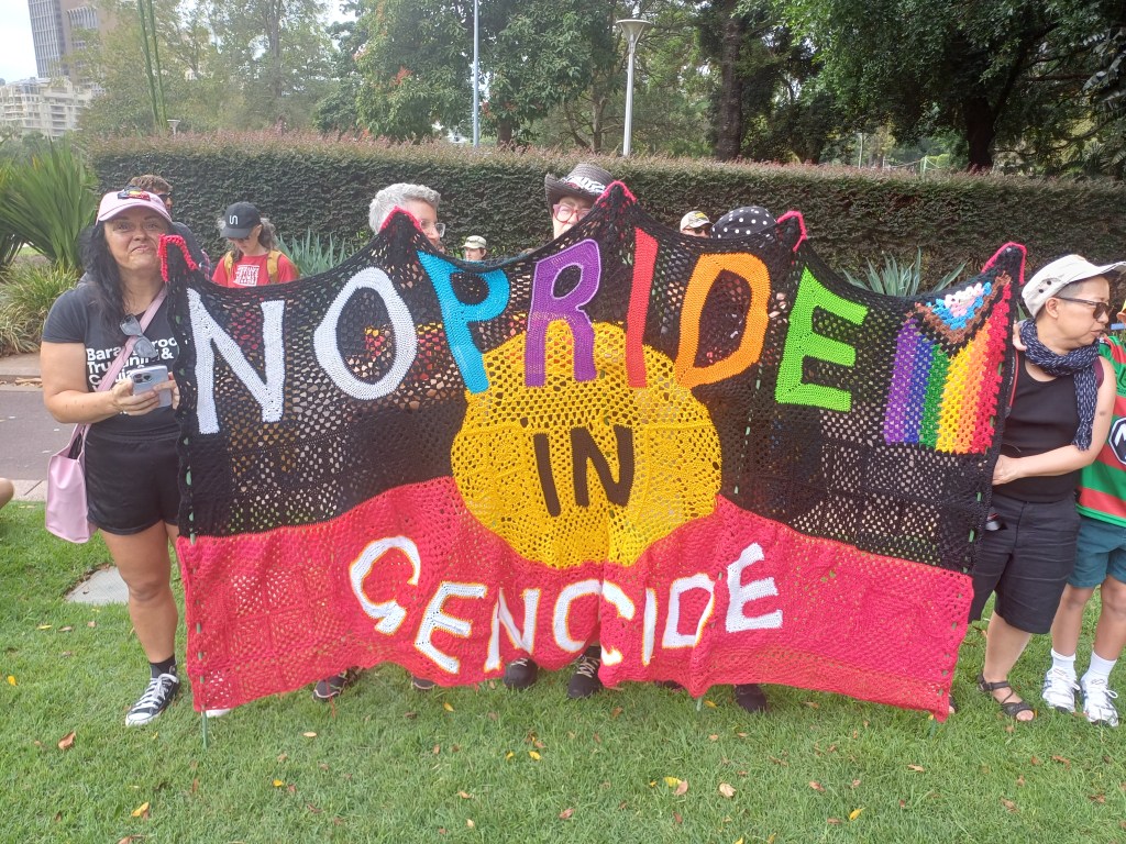 Large crocheted Australian Aboriginal flag (black sky, red earth, yellow sun in the middle) with the words 'NO PRIDE IN GENOCIDE' across it, also in crochet. For the word 'pride', each letter is in a different rainbow colour, and there is also a small progress pride flag attached.
